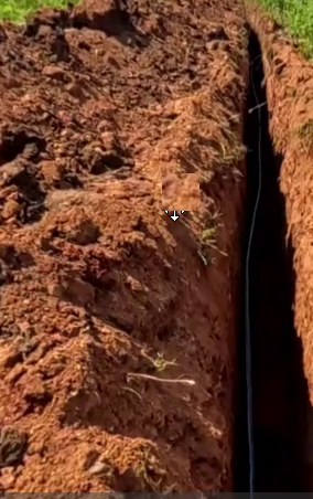 An open fibre trench cut through red soil with the duct lying in the bottom, photographed before backfill