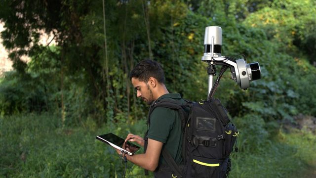 A surveyor walking through vegetation carrying the backpack lidar unit, operating a tablet as he goes